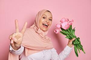 magnifique arabe musulman femme portant rose hijab, en portant une bouquet de tulipes, montrant paix main signe, souriant à caméra photo