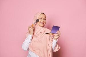 attrayant milieu vieilli arabe musulman femme dans rose hijab, appliquant maquillage sur sa affronter, isolé sur rose Couleur Contexte photo