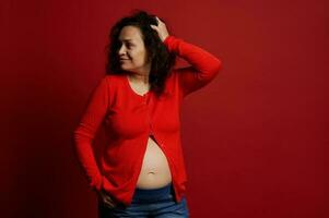 portrait de multi ethnique mature Enceinte femme dans rouge chemise, à la recherche de côté, posant nu ventre sur isolé rouge Contexte photo