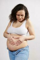 Enceinte femme dans blanc sous-vêtements et denim jeans, en mettant sa mains sur sa ventre, fabrication cœur forme, isolé sur blanc photo