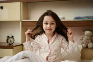 charmant peu fille veille en haut dans matin, redressage sa magnifique longue cheveux, souriant à caméra, tandis que séance sur lit photo