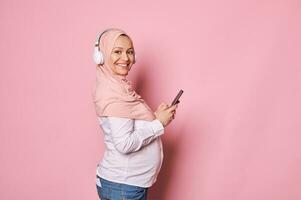 souriant content Enceinte musulman femme dans rose hijab et sans fil écouteurs, vérification Nouveau mobile application sur téléphone intelligent photo