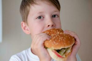 fermer portrait de une peu garçon en mangeant une énorme Burger, sur une blanc Contexte photo