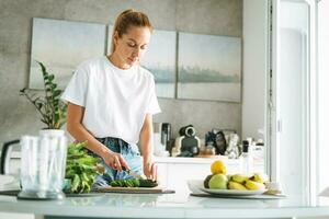 Jeune svelte femme dans blanc T-shirt et bleu jeans cuisine en bonne santé nourriture avec concombre dans cuisine à Accueil photo