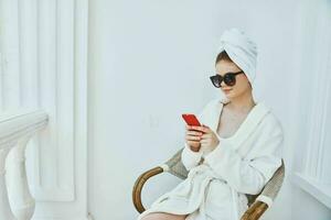 une femme dans une blanc peignoir est assis dans une chaise et les usages une téléphone avec des lunettes de soleil dans le Matin photo