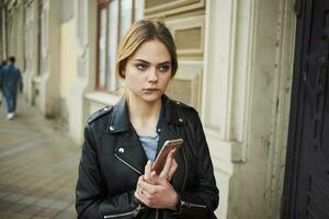 femme sur le rue avec une téléphone dans sa mains cuir veste marcher près le bâtiment photo