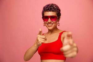 mode portrait de une femme avec une court la Coupe de cheveux dans coloré des lunettes de soleil avec inhabituel accessoires avec des boucles d'oreilles sourit sur une rose brillant Contexte spectacles sa mains à le caméra photo