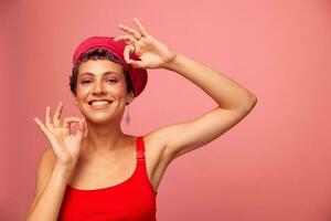 Jeune athlétique femme avec une court la Coupe de cheveux et violet cheveux dans une rouge Haut et une rose chapeau avec un athlétique figure sourit et grimaces à la recherche à le caméra sur une rose Contexte photo