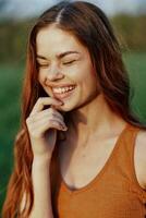 fermer portrait de une souriant Jeune femme avec yeux fermé dans le Soleil avec rouge cheveux dans la nature photo