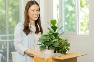 content asiatique Jeune femme, client reçu, déballer paquet carton ouvert parcelle dans papier carton boîte, en portant céramique plante pot. en mouvement, en train de préparer dans Nouveau loger, déménagement. en ligne magasin livraison à maison. photo