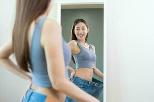 forme mince, mince taille, attrayant svelte asiatique Jeune femme, magnifique fille main spectacle forme sa poids perte, portant dans grand, grand ou plus de Taille jeans et à la recherche dans miroir. gens corps en forme en bonne santé. photo