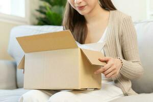 content asiatique Jeune femme, fille client séance sur canapé à maison, main dans ouverture et déballage papier carton boîte carton parcelle après achat commande cadeau, achats en ligne, livraison un service concept. photo