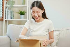 content excité, asiatique Jeune femme, fille client séance sur canapé à maison, ouverture et déballage papier carton boîte carton parcelle après achat commande cadeau, achats en ligne, livraison un service concept. photo