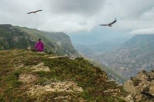 femme est assis avec sa retour à le montagnes au dessus une falaise, aigles en volant dans le spectaculaire ciel. photo