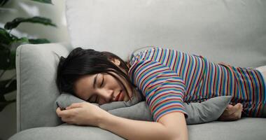 un épuisé Jeune asiatique femelle mensonge sur une confortable canapé dans le vivant pièce en train de dormir après travailleur journée photo