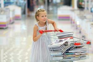 le enfant a rempli une panier de livres. peu fille dans le livre magasin. vente. enfant dans le bibliothèque avec une chariot de livres. photo