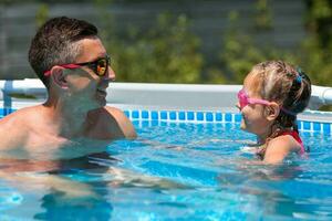 père dans des lunettes de soleil nage dans le bassin avec le sien fille. papa et peu fille dans nager des lunettes de protection nager dans le bassin. une content homme et une enfant dans le l'eau. photo