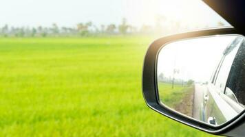 vue de à côté de miroir aile de voiture pouvez vue voitures derrière dans le distance sur asphalte route. Contexte image est une vert jaunâtre riz champ dans le Matin. photo