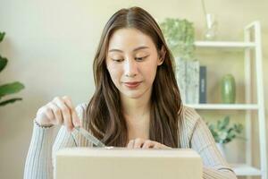 content asiatique Jeune femme, fille client séance sur canapé à maison, main dans ouverture et déballage papier carton boîte carton parcelle après achat commande cadeau, achats en ligne, livraison un service concept. photo