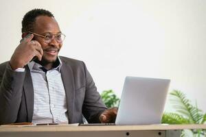 un africain américain homme d'affaire dans une noir costume les usages une mobile téléphone à communiquer avec clients et une portable à vue entreprise bénéfices. à construire confiance pour les clients à viens et investir dans le Bureau photo