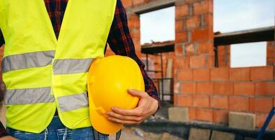 construction ouvrier sur site zone. ouvrier dans Jaune gilet en portant difficile chapeau et rester dans de face de le bâtiment zone. construction ouvrier sur le travail zone. photo