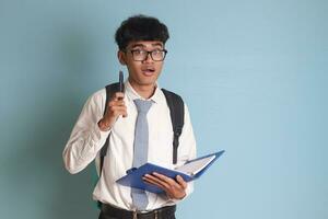 indonésien Sénior haute école étudiant portant blanc chemise uniforme avec gris attacher l'écriture sur Remarque livre en utilisant stylo. isolé image sur bleu Contexte photo