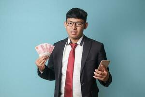portrait de Jeune asiatique affaires homme dans décontractée costume à la recherche à caméra tandis que en portant téléphone mille roupies. isolé image sur bleu Contexte photo