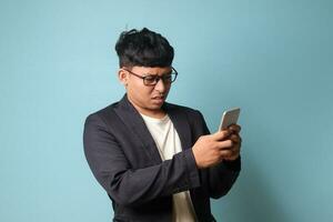 portrait de Jeune asiatique affaires homme dans décontractée costume dans incrédulité tandis que à la recherche à téléphone. isolé image sur bleu Contexte photo
