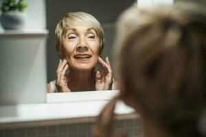 mature femme à la recherche à sa visage dans le salle de bains. elle est examiner les rides sur sa peau. photo