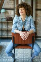 sérieux Jeune femme portant denim veste séance sur cuir chaise photo