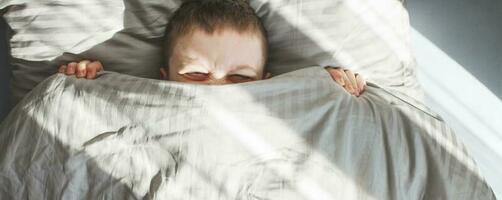 garçon sommeil dans le lit. enfant mensonges sur oreiller et couvertures le sien visage avec une couverture. bannière photo