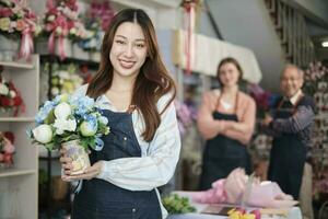 magnifique Jeune asiatique femelle fleuriste ouvrier avec vase de fleur sourit et regards à caméra dans de face de collègues équipe dans coloré fleur boutique, petit affaires profession, content pme entrepreneur. photo