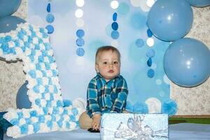 une magnifique bébé est célébrer le sien premier anniversaire. portrait de le bébé. une garçon dans une bleu chemise parmi des ballons et cadeaux. photo