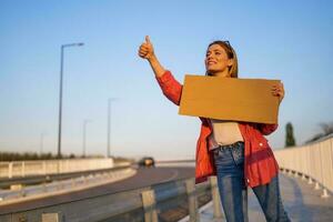 femme est auto-stop sur bord de la route en essayant à Arrêtez auto. elle est en portant Vide papier carton pour votre texte. photo