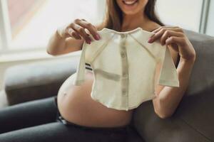 Enceinte femme relaxant à maison. elle est séance sur lit et en portant bébé vêtements. photo