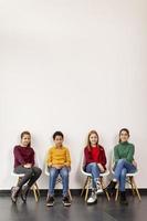 Portrait de mignons petits enfants en jeans assis sur des chaises contre le mur blanc photo