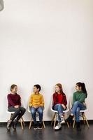 Portrait de mignons petits enfants en jeans assis sur des chaises contre le mur blanc photo