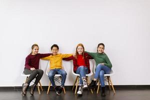 Portrait de mignons petits enfants en jeans assis sur des chaises contre le mur blanc photo