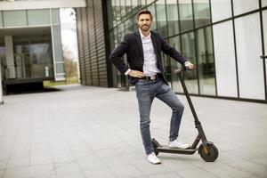 Jeune homme d'affaires dans un vêtement décontracté debout sur un scooter électrique par un immeuble de bureaux lors d'une réunion d'affaires photo