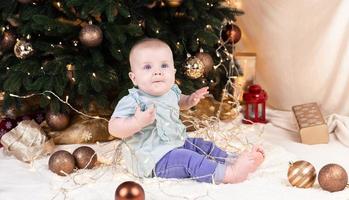 un bébé en jean est assis près d'un arbre de Noël et joue avec une guirlande dans laquelle il est empêtré photo