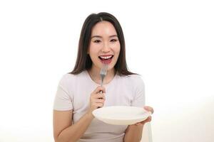 magnifique Jeune Sud est asiatique femme faire semblant agissant posant en portant vide fourchette cuillère blanc assiette dans main manger goût Regardez voir blanc Contexte sourire content nourriture dans bouche photo