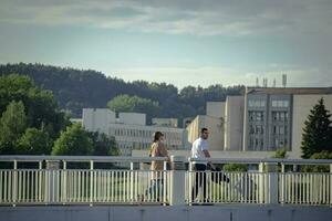 Vilnius, Lituanie 03 06 2022 Jeune femme un homme en marchant sur une blanc pont avec vert des arbres et Bureau bâtiment sur Contexte photo