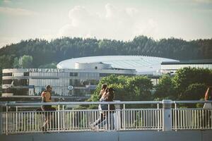 Vilnius, Lituanie 03 06 2022 Jeune gens et Hommes en marchant sur une blanc pont plus de rivière avec vert des arbres et Bureau bâtiment sur Contexte photo