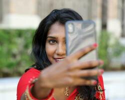 Jeune attrayant magnifique Indien femme dans rouge et noir sari prise selfie soi portrait en utilisant intelligent téléphone en plein air Urbain brique mur Contexte photo