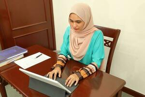 Jeune asiatique malais musulman femme portant foulard à Accueil Bureau étudiant asseoir à table ordinateur livre fichier papier étude travail photo