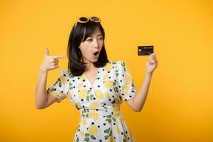 portrait Jeune asiatique femme content souriant dans printemps robe montrant Plastique crédit carte isolé sur Jaune Contexte. payer, argent et achat achats Paiement concept. photo