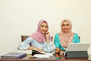 deux Jeune asiatique malais musulman femme portant foulard à Accueil Bureau étudiant séance à table parler mêler Regardez à ordinateur livre document étude discuter sourire content photo