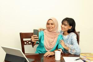 deux Jeune asiatique malais musulman femme portant foulard à Accueil Bureau étudiant séance à table téléphone ordinateur livre document selfie soi portrait avec téléphone intelligent photo
