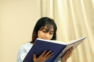 Jeune asiatique malais musulman femme portant baju kurung robe à Accueil asseoir du repos sur canapé lis livre Remarques visage derrière le livre photo