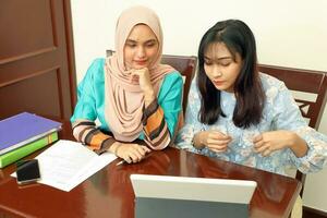 deux Jeune asiatique malais musulman femme portant foulard à Accueil Bureau étudiant séance à table parler mêler Regardez à ordinateur livre document étude discuter lis type écrire sourire content photo